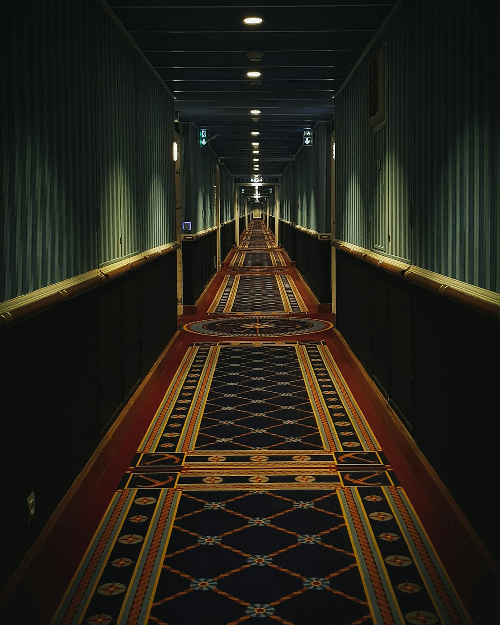 A long grand-hotel corridor, patterned carpet running to a lit door.