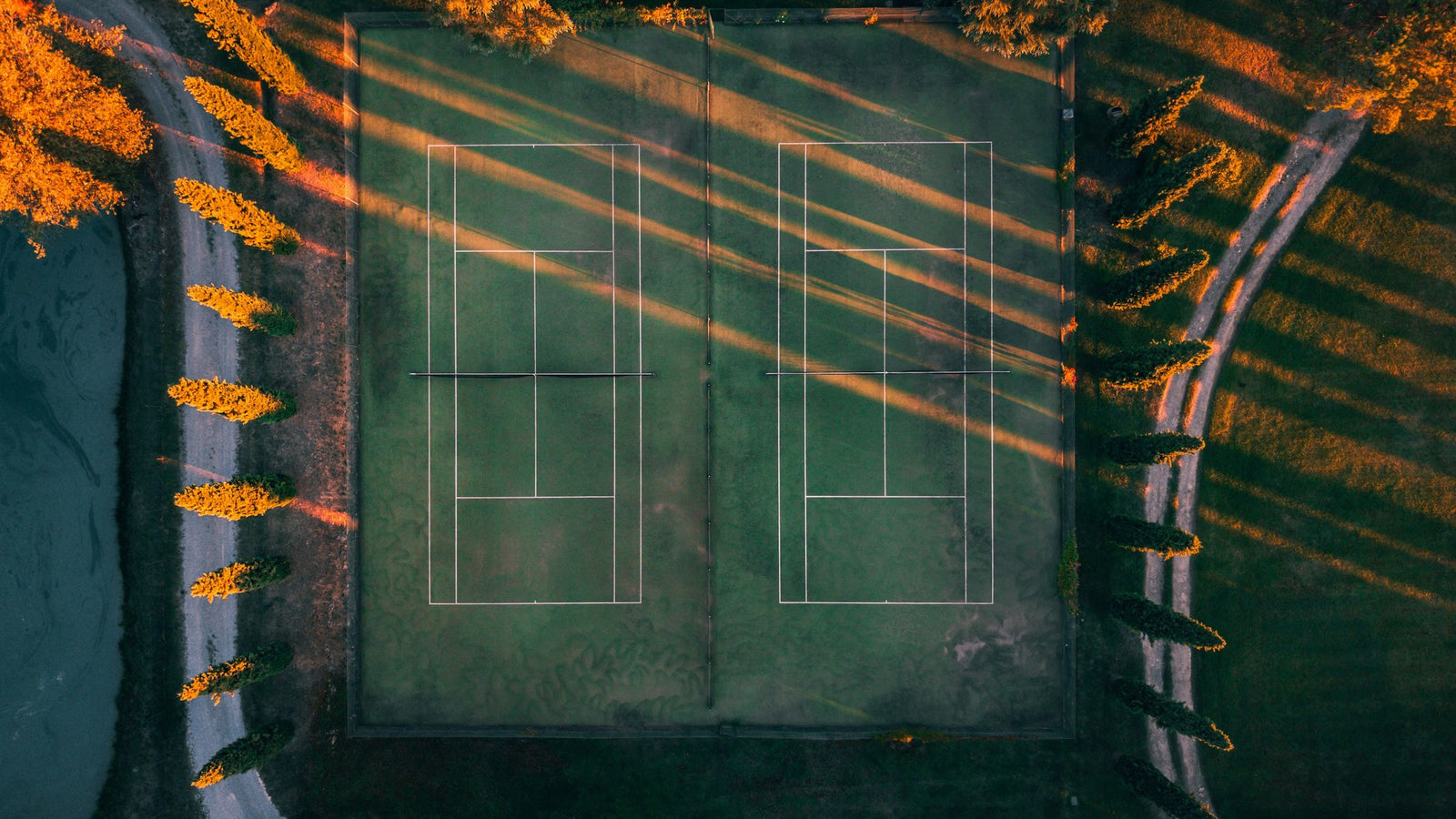 A grass tennis court seen from above at sunset, long cypress shadows across the lines.